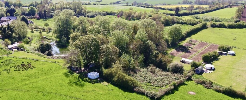 Aerial view of a lush green landscape with several Mongolian yurts set amidst trees and a small pond, showcasing a serene outdoor retreat.