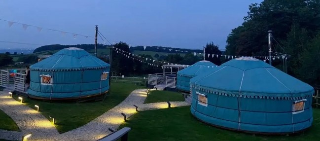 A picturesque view of multiple handcrafted Mongolian yurts illuminated at dusk, surrounded by grassy fields and a gravel pathway. 