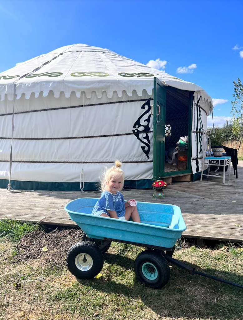 A young child sitting in a blue wagon in front of a large glamping yurt