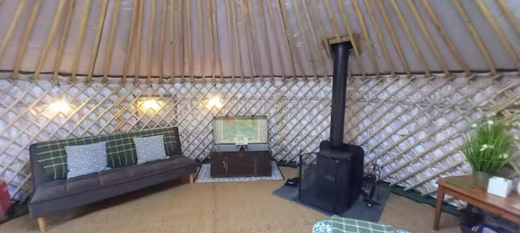 Luxurey glamping yurt interior seating - Visit our Oxford Show yurt or Speak to Us Interior of a luxury glamping yurt featuring a grey sofa, wood-burning stove, and traditional wooden lattice walls.
