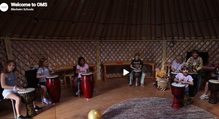 A group of children and teacher participating in drumming circle inside a spacious, lattice - walled wooden yurt with a circular rug and natural lighting.
