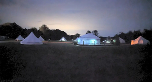 Luxury bell tents yurt night view - Welcome to Yurt Specialists a row of illuminated whit bell tents and a centra yurt glowing at night on a dark green field under a twilight sky