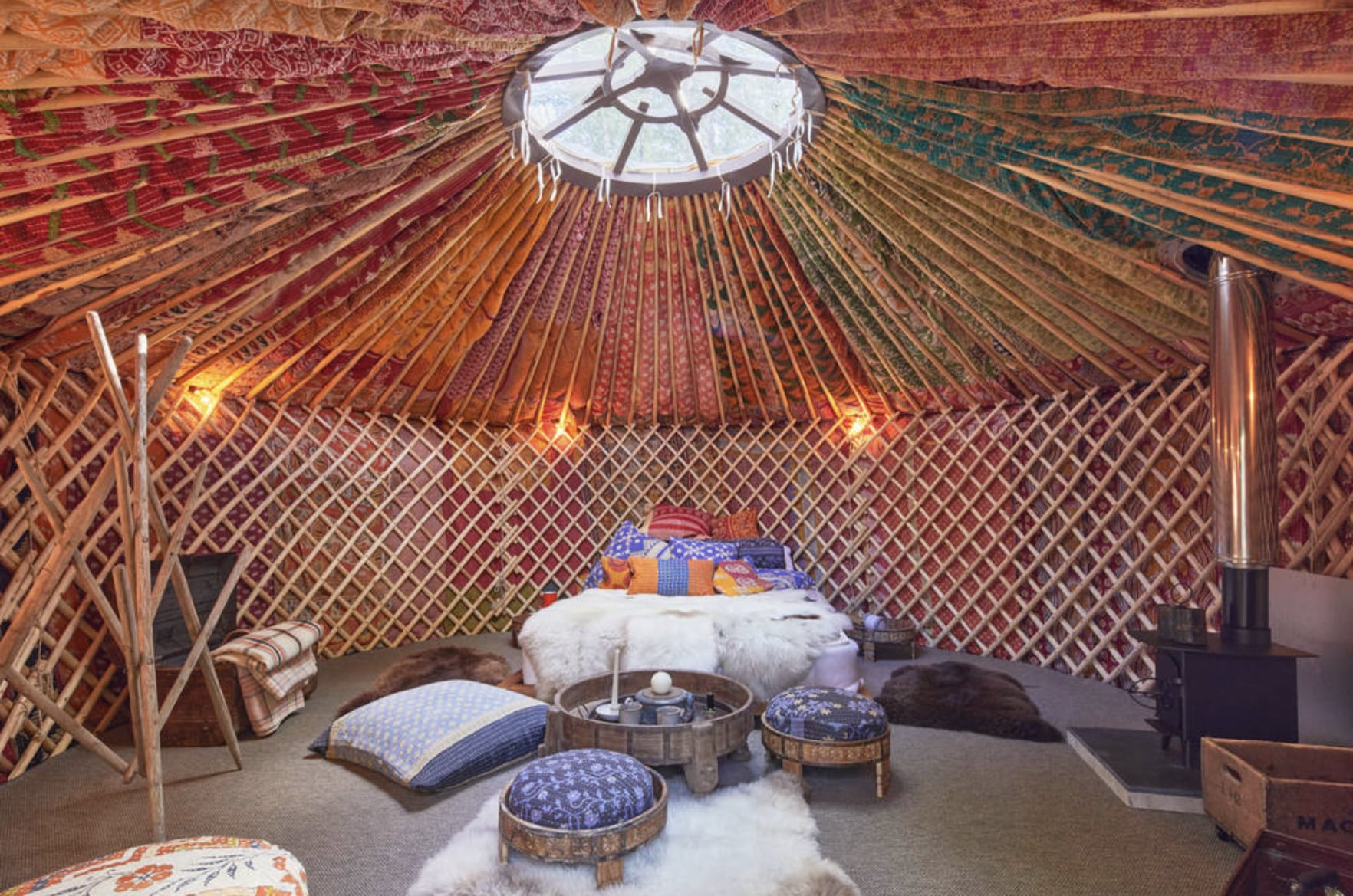 A wide-angle view of a luxurious glamping yurt interior, featuring rich, colorful patterned textiles draped across the ceiling, handcrafted wooden lattice walls, and a central skylight (toono). The cozy space includes a large, plush bed with colorful pillows, multiple fur rugs, low-profile seating, a multi-legged decorative pole, and a functional wood-burning stove with a polished chimney pipe. Warm, ambient lighting is present.