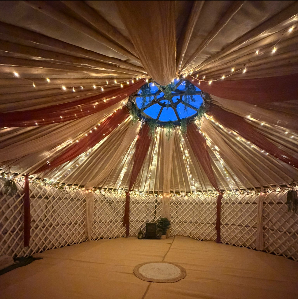 A wide-angle interior view of a traditional circular yurt decorated for a wellness retreat. The ceiling features draped cream and terracotta fabric radiating from a central blue skylight, accented by strings of warm fairy lights. The wall consists of a light wood lattice frame, and the floor is covered in tan mats with a small circular rug in the centre.