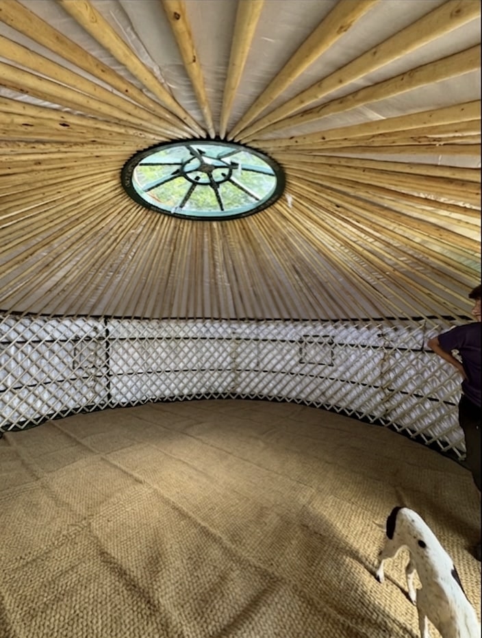 A interval of a traditional Mongolian yurt, featuring wooden lactic wall, radiant ceiling poles, and a beautiful skylight. The floor is covered in natural matting ready for children to learn and play.