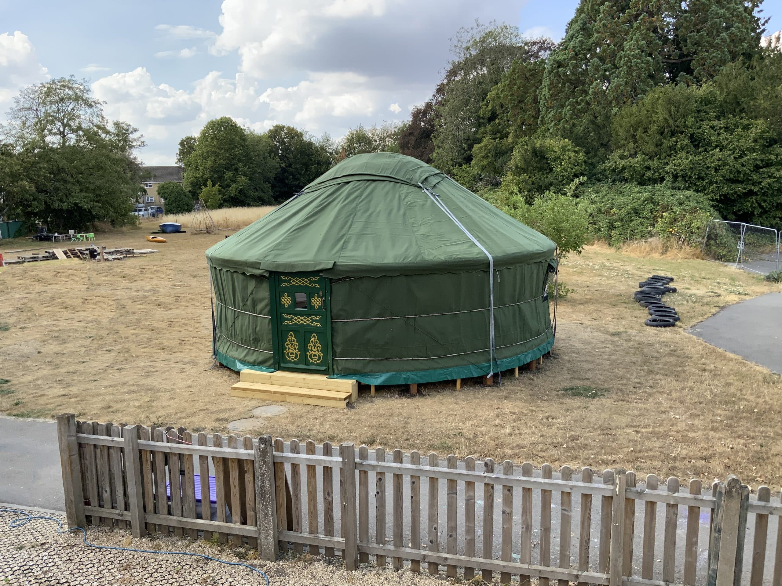 Green traditional Mongolian yurt with decorative wooden door pitched in an outdoors school ground surrounded by trees and playground equipment.