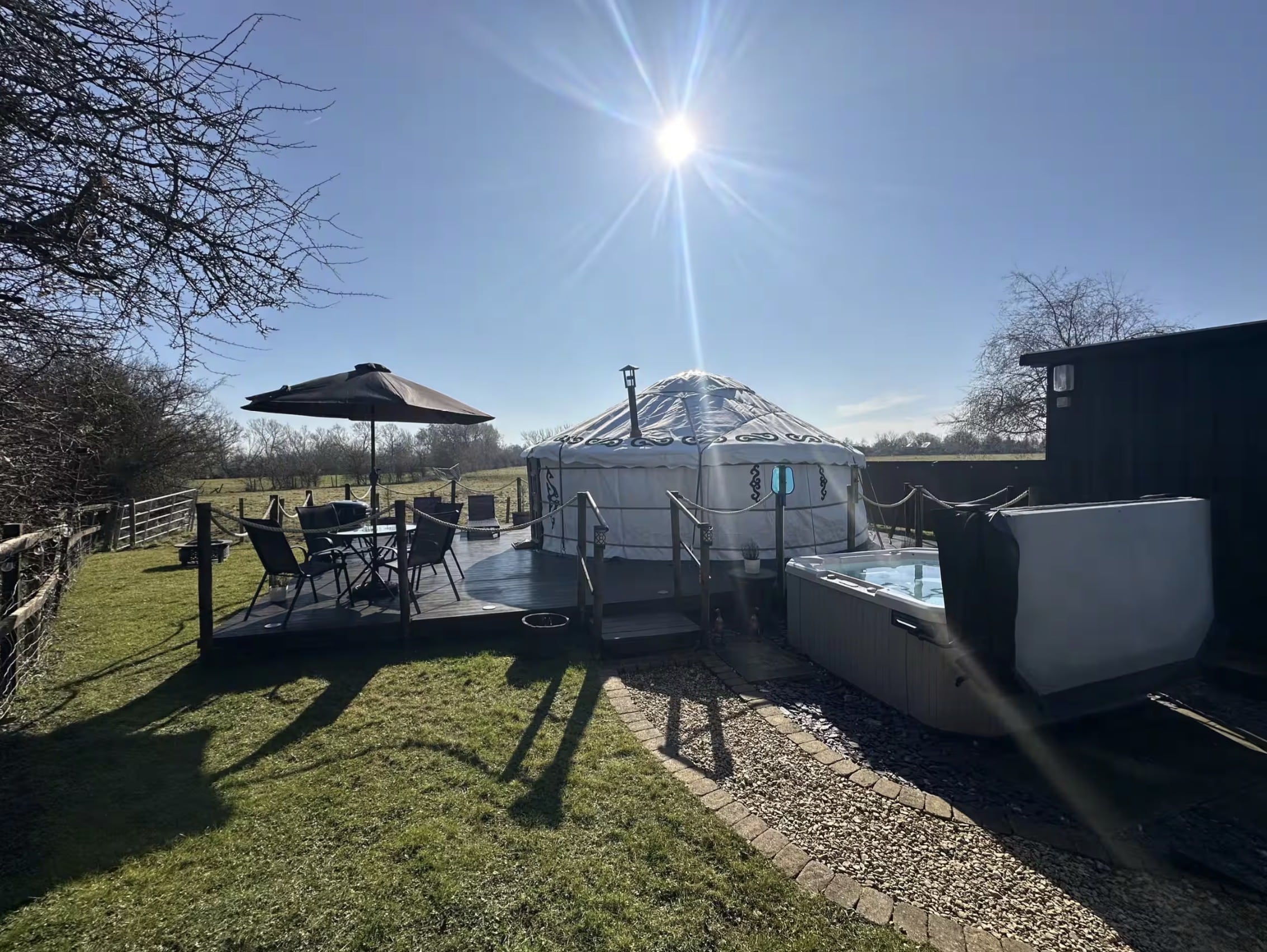 White canvas glamping yurt set on a wooden decking with outdoor patio seating, umbrella, and a hot tub under a sunny sky.